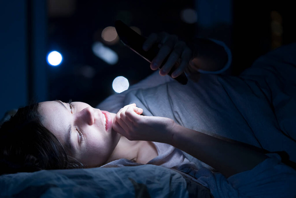 Young girl in a bed with a phone at night. - 写真・画像