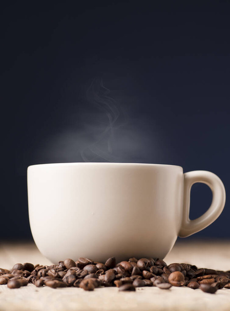 weiße Tasse Kaffee mit Rauch auf rustikalem Holztisch, mit gerösteten Kaffeebohnen, blauer Hintergrund - Foto, Bild