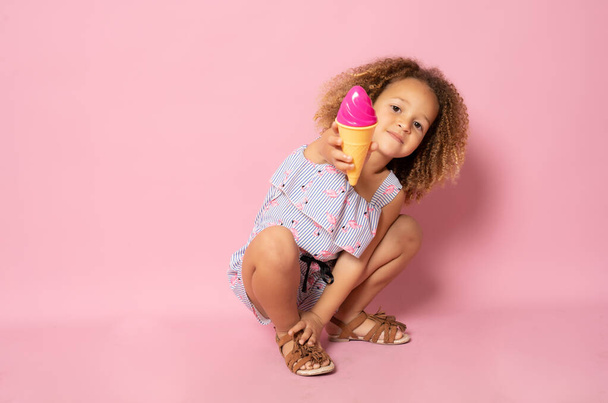 Милая маленькая девочка в летней одежде с мороженым сидя на полу изолированы на розовом фоне. - Фото, изображение