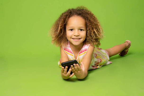 Linda niña en vestido de verano acostado en el suelo usando el teléfono móvil sobre fondo verde. - Foto, Imagen