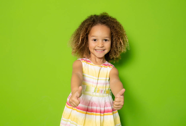 Söpö pieni tyttö kesällä mekko osoittaa peukalot pystyssä eristetty yli vihreä tausta. - Valokuva, kuva