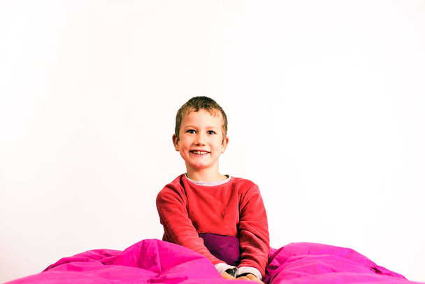 Handsome little boy wakes up and poses sitting on his bed covered by a duvet, isolated on white studio background. - Φωτογραφία, εικόνα