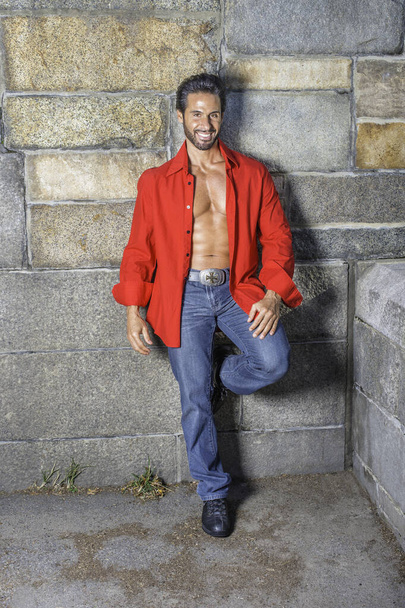 Dressing in a red long sleeve shirt, unbuttoned, blue jeans, leather shoes, a sexy, middle age guy with mustache and beard is leaning against the wall, smilingly looking at you - Fotó, kép