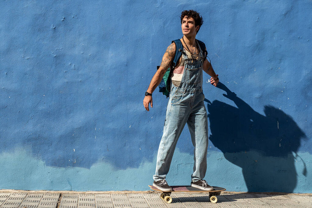 Stylish spanish guy hipster skateboarding on waveboard. City lifestyle. Blue wall. A lot of copy space. Hobby. - Photo, Image