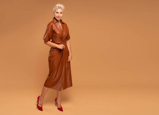 Beautiful blonde middle aged woman with short hairstyle posing in brown medium dress. Fashion photo. Elegant businesswoman. Full photo. A lot of copy space - Foto, immagini
