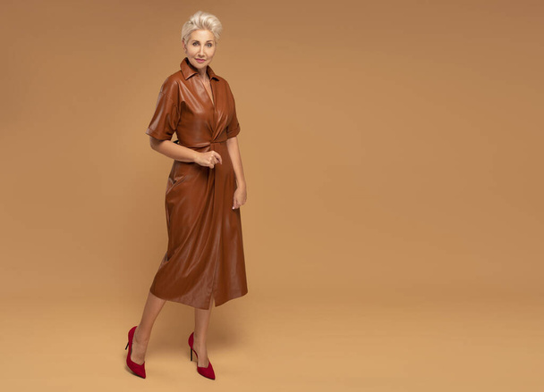 Beautiful blonde middle aged woman with short hairstyle posing in brown medium dress. Fashion photo. Elegant businesswoman. Full photo. A lot of copy space - Фото, изображение