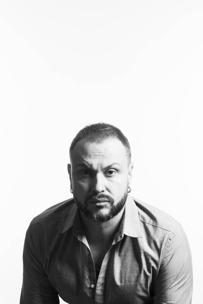 Emotive portrait of bearded charismatic muscular 40 years old man sitting over white background, looking at camera. Short haircut. Black and white studio shot. Text space - Valokuva, kuva