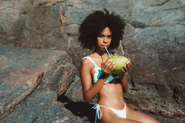 A portrait of a sensual young African-American lady with curly Afro hair, sitting on the rock on the beach of a tropical resort on a sunny day, and drinking a fresh coconut water from the coco - Foto, immagini