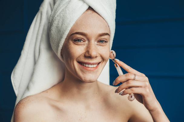 Caucasian freckled woman wrapped in towels massaging face with jade roller. Close up portrait. Smiling face. Skin care. Beauty face. - Фото, зображення
