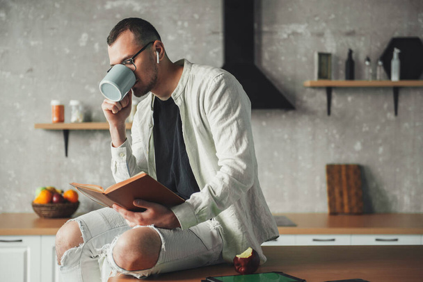Portret van een man die koffie drinkt met een bril op en een boek leest terwijl hij in de keuken rust. Koffiepauze. - Foto, afbeelding