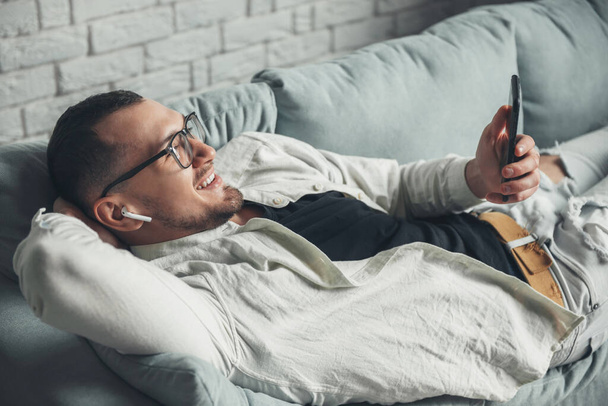 携帯電話を持って眼鏡をかけ、ソファに横になって笑っている男。エアポッドだ。無線通信だ。モバイル通信. - 写真・画像