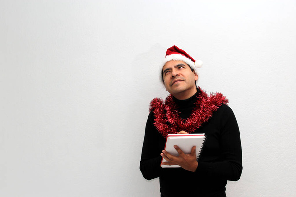 Взрослый латиноамериканец в шляпе и рождественской гирлянде показывает баннер и пустой блокнот, чтобы оставить сообщение поздравления, любовь или список желаний - Фото, изображение