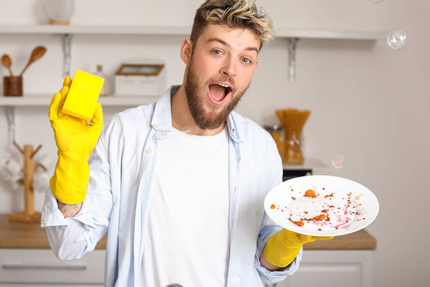 Surprised young man with sponge and dirty plate in kitchen - Фото, изображение