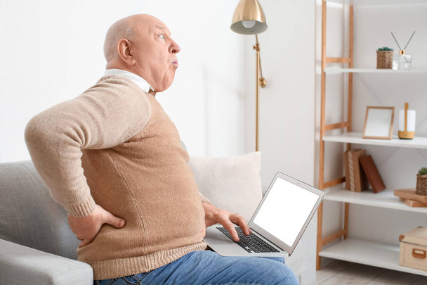 Senior man met laptop die last heeft van rugpijn op de bank thuis - Foto, afbeelding