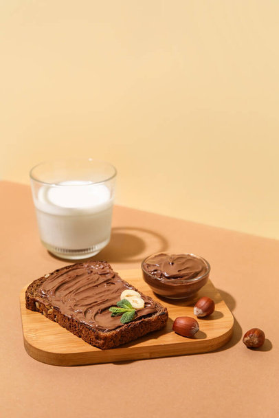 Leipä suklaatahnalla, hasselpähkinöillä ja lasillisella maitoa beigellä pohjalla - Valokuva, kuva