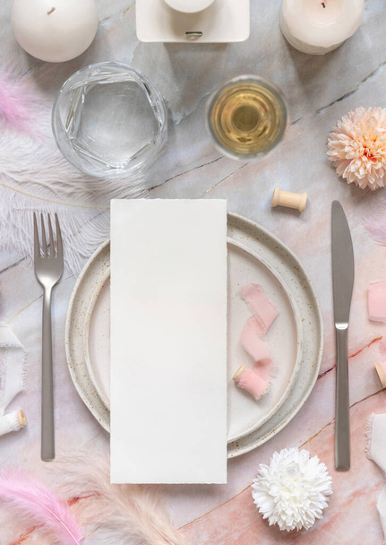 Свадебный стол с бумажной карточкой на фарфоровых тарелках рядом с розовыми цветами, перьями и шелковыми лентами меню макет. Соблазн с вертикальной карточкой лежал плоским слоем. Макет пригласительной или меню карты, копировальная площадка - Фото, изображение