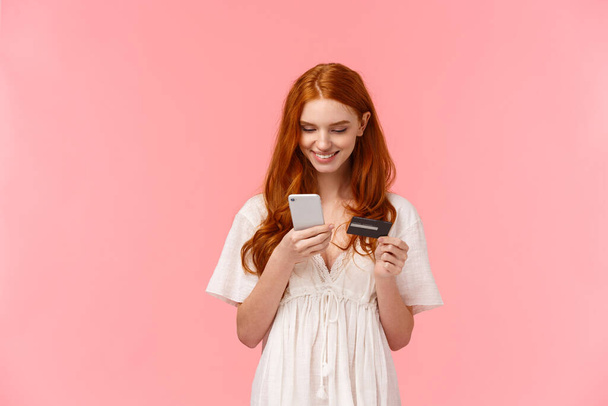 Vyötärö-up muotokuva karismaattinen söpö punapää nainen valkoisessa mekossa, aseta hänen luottokorttinumeronsa älypuhelimeen, ostaa sovellus, tehdä online-tilauksen, ostokset verkkokaupassa, hymyillen esillä - Valokuva, kuva
