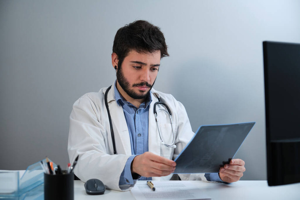 Giovane medico che guarda una radiografia di un bacino e colonna vertebrale alla scrivania. - Foto, immagini