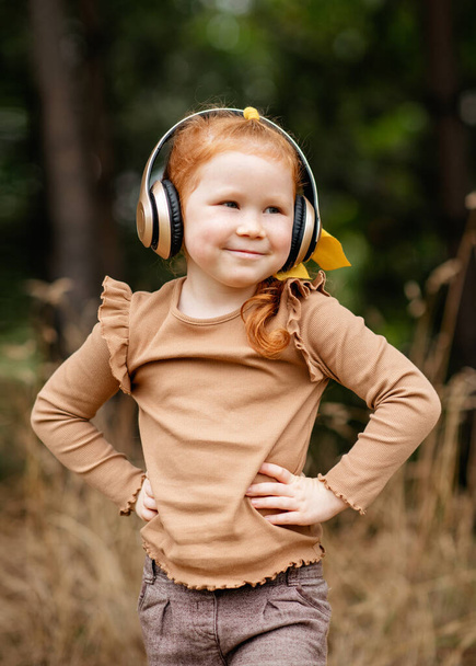 malá veselá dívka s rudými vlasy a sluchátky, která poslouchá hudbu v parku na podzim - Fotografie, Obrázek