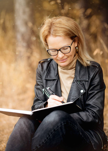 a smiling woman in a black leather jacket and glasses  sitting in the park and drawing on autumn day - Foto, Bild