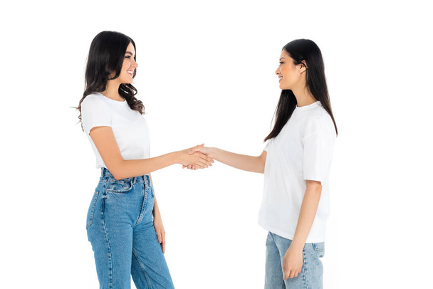 happy brunette multiethnic women looking at each other and shaking hands isolated on white - Фото, изображение