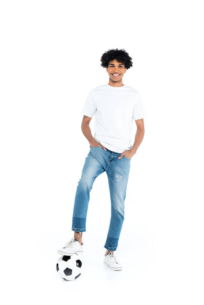 smiling african american man standing with hands in pockets of jeans near soccer ball on white - Foto, afbeelding