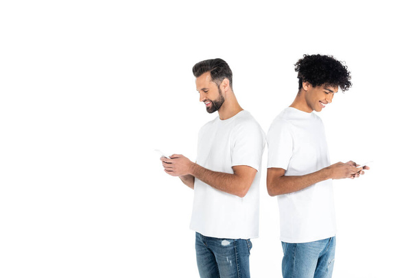 smiling interracial men messaging on mobile phones while standing back to back isolated on white - Фото, зображення
