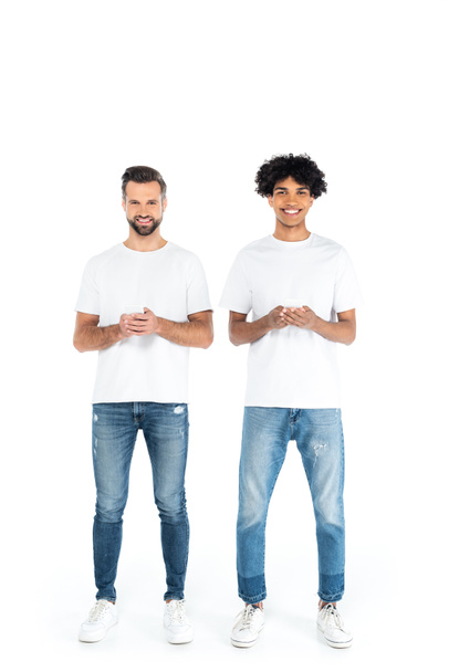 full length view of happy multiethnic men in t-shirts and jeans chatting on smartphones on white - Foto, imagen