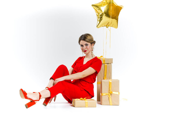 Cheerful attractive woman in red costume having fun sitting on big gift box isolated on white background and gold stars. Celebrating birthday, anniversary. Shopping for holiday concept, saleout - Photo, Image