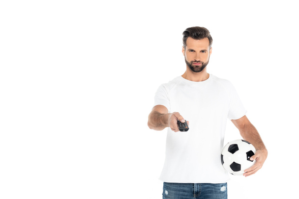 bearded man in t-shirt clicking tv channels on remote controller while holding soccer ball isolated on white - 写真・画像