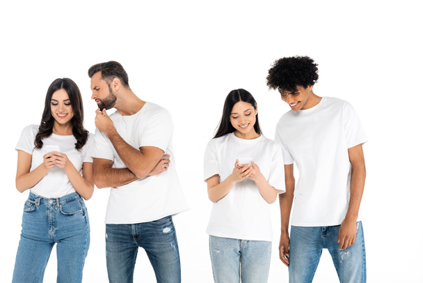 веселі міжрасові жінки обмінюються повідомленнями на мобільних телефонах поблизу цікавих чоловіків ізольовані на білому
 - Фото, зображення