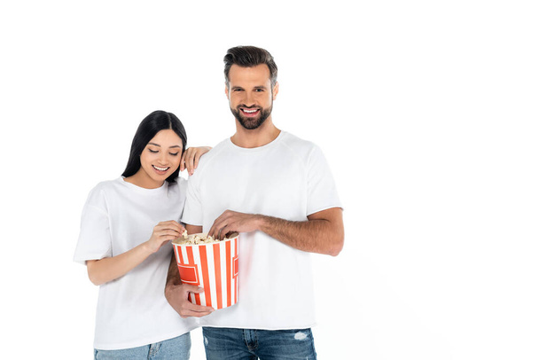 smiling man holding popcorn bucket near happy asian woman isolated on white - Foto, afbeelding