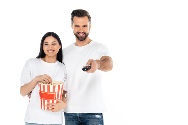 tevreden man klikken tv kanalen in de buurt van aziatische vrouw met emmer van popcorn geïsoleerd op wit - Foto, afbeelding