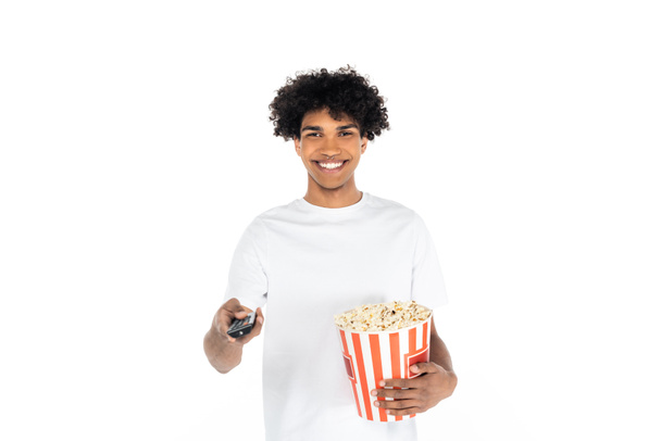 homme afro-américain joyeux avec un seau de pop-corn en cliquant sur les chaînes de télévision isolées sur blanc - Photo, image