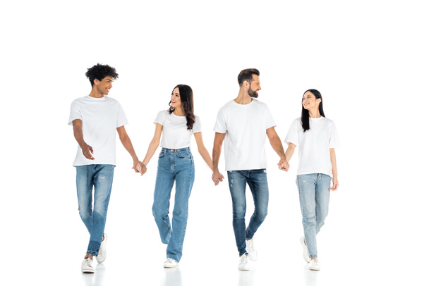 full length view of interracial friends in jeans looking at each other and holding hands while walking on white - Photo, Image