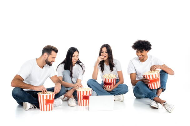 smiling multiethnic friends eating popcorn while watching movie on laptop on white - Valokuva, kuva