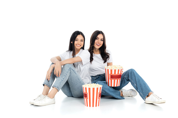 brunette interraciale vrouwen glimlachen op camera terwijl zitten in de buurt van grote emmers van popcorn op wit - Foto, afbeelding