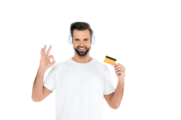 pleased man in headphones holding credit card and showing okay gesture isolated on white - Fotó, kép