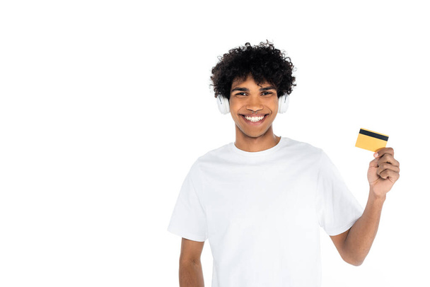 jovem afro-americano em fones de ouvido segurando cartão de crédito e sorrindo para a câmera isolada em branco - Foto, Imagem