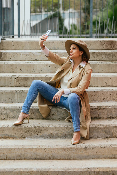 Een elegante volwassen vrouw zittend op stappen buiten en het nemen van een selfie - Foto, afbeelding