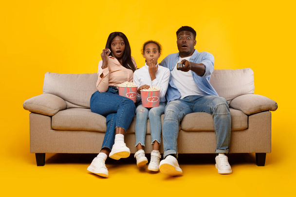 Sorprendido joven familia viendo la televisión con palomitas de maíz sentado en el sofá - Foto, imagen