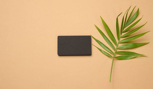 a stack of rectangular black paper business cards and a leaf of a palm tree on a brown background. View from above, modern template for branding identity - Foto, afbeelding