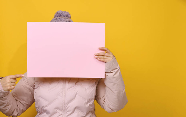 Frau in pinkfarbener Winterjacke und Hut hält leeres rosafarbenes Blatt Papier auf gelbem Hintergrund. Saisonverkauf. Platz für die Inschrift - Foto, Bild