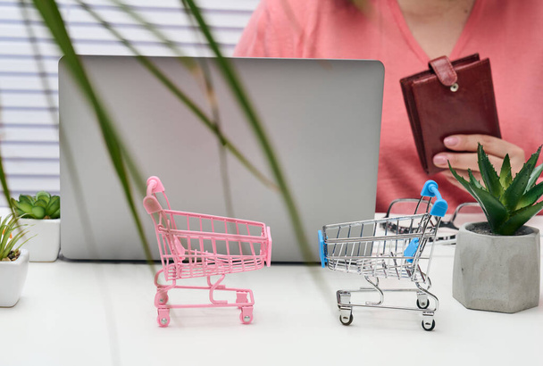 two empty miniature carts, an open laptop and a woman with a purse sitting at a white table. Online shopping, sale - Fotó, kép