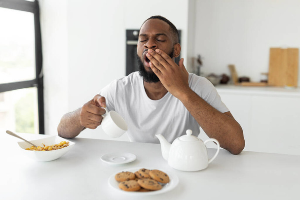 Black man yawning, pouring coffee away from cup - Foto, afbeelding