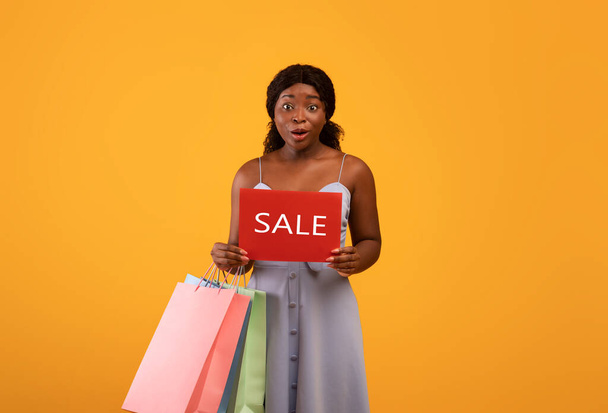 Surprised millennial black woman holding shopping bags and SALE sign over orange studio background - Foto, Bild