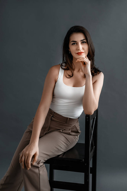 Carefree young woman in smart casual wear sitting on a chair, posing in studio over gray background. Relaxed and beautiful young woman - Zdjęcie, obraz
