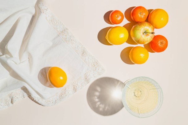 Creative composition made of glass with fruit juice, lemon, mandarin and apples on pastel beige sunlit background with white cloth and shadows. Summer and refreshment concept. Harvest theme - Fotó, kép