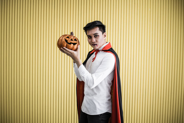 Halloween concept of young Asian man in costume dracula and hold Halloween orange pumpkin on yellow background - Фото, изображение