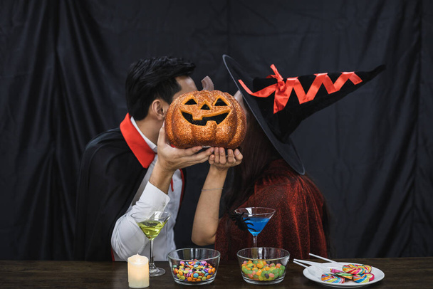 Młoda azjatycka para w kostiumie czarownica i dracula z okazji Halloween party i pocałował z dyni zakrywającej twarz. Para w kostiumie świętować Halloween strona czarny tkanina tło. - Zdjęcie, obraz